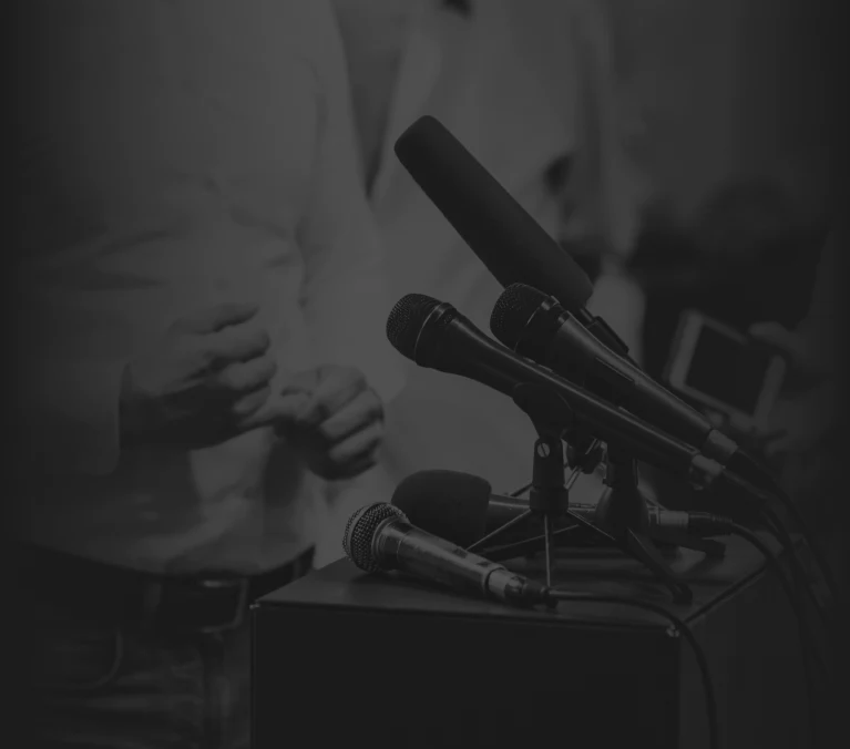 Black and white image of a person standing near a podium with multiple microphones. The person appears to be preparing for a speech or addressing an audience, with their hands visible but not their face.