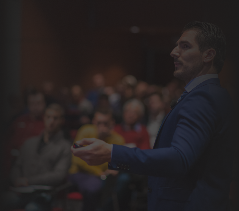 A man delivering a public speech in a formal setting, wearing a suit and addressing an attentive audience. The background shows a room filled with people focused on his presentation, emphasizing his confident and professional demeanor while engaging with the crowd.